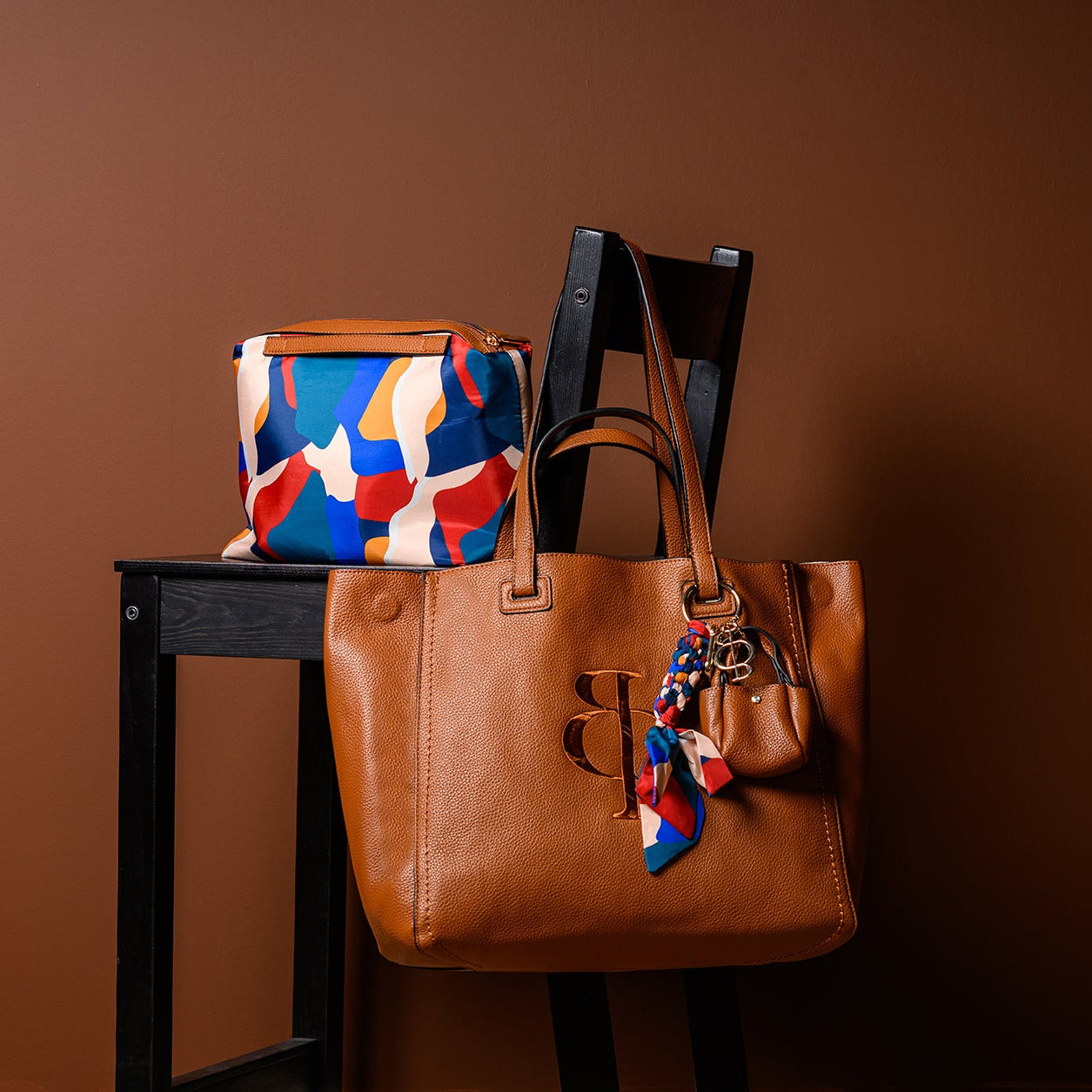 Brown handbag with a colorful scarf draped over it on a wooden stool against a brown background