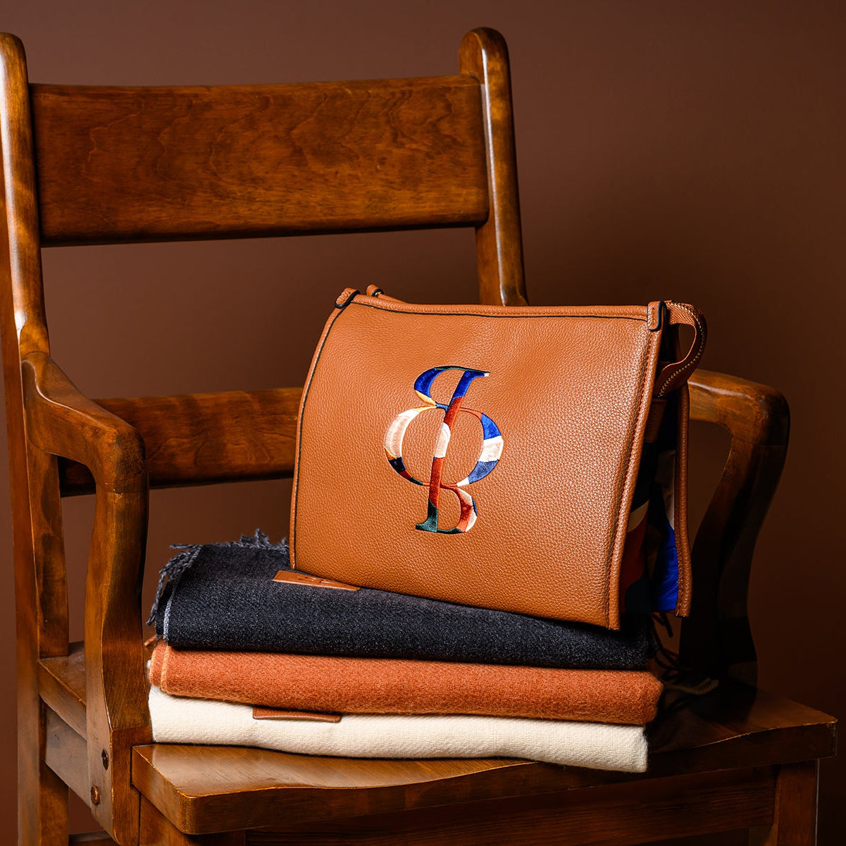 Brown leather bag with a logo on top of folded cloths on a wooden chair against a brown background